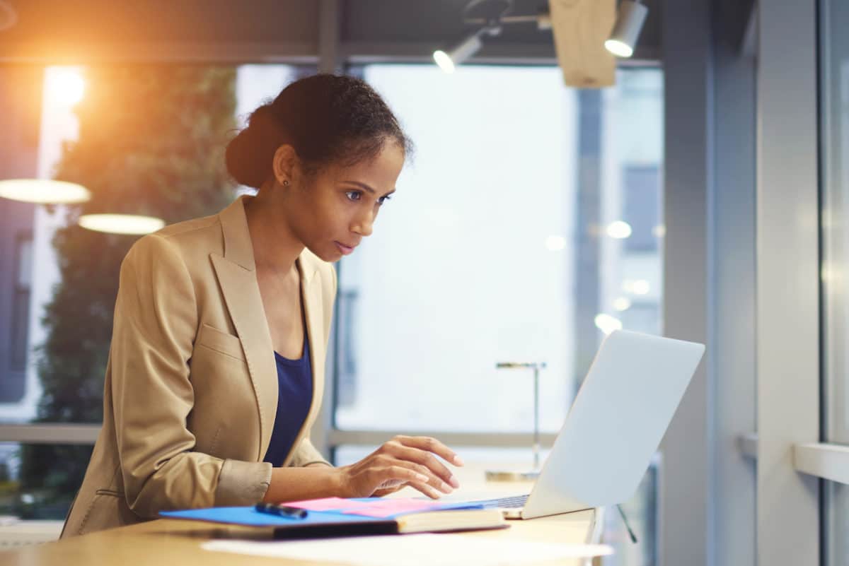 A woman looks intently on her laptop while papers sit next to her on the desk