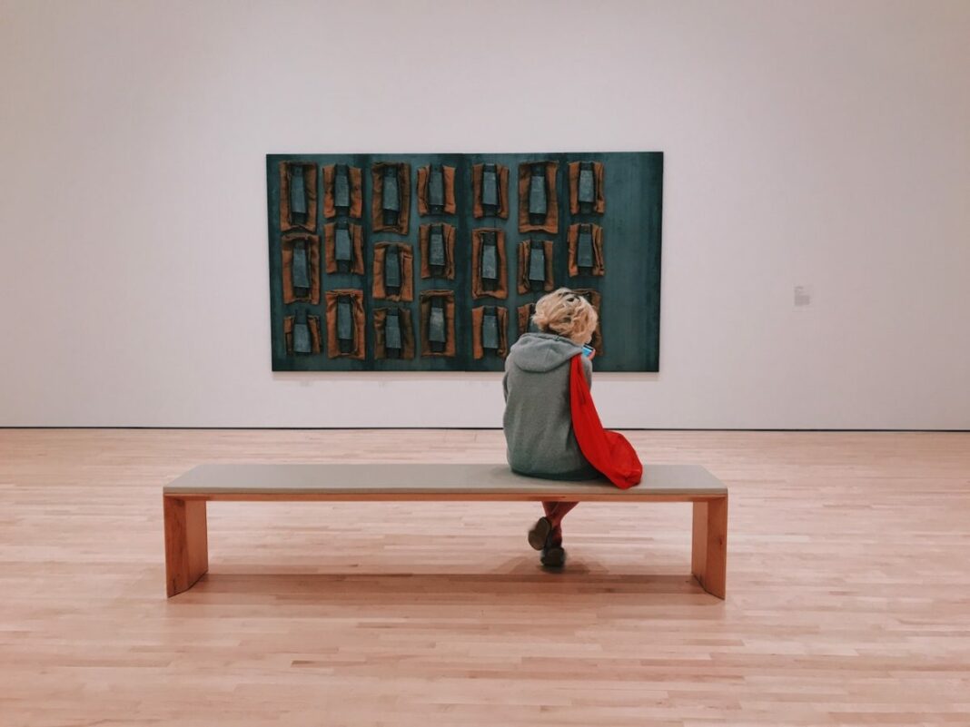 woman sitting on bench inside museum room with one painting on the wall